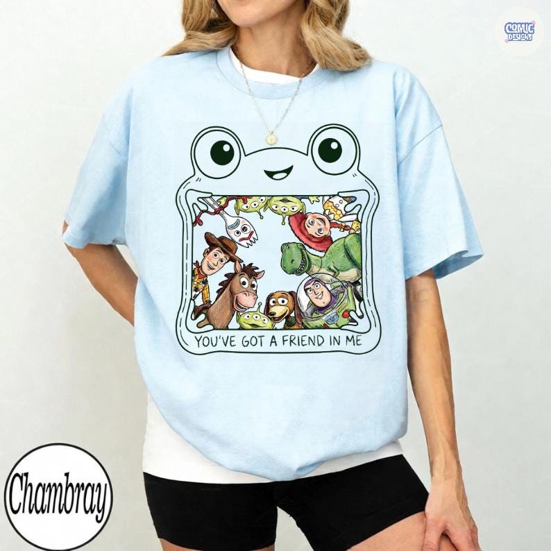 May include: A light blue t-shirt featuring a graphic of a frog-shaped frame containing various animated characters and the text "YOU'VE GOT A FRIEND IN ME". The design includes illustrations of a cowboy, a space ranger, a dinosaur, and other figures.