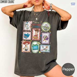May include: A charcoal gray short-sleeve t-shirt featuring a graphic print of various Toy Story characters in postage stamp-style frames. The text "You've Got A Friend In Me" is printed in the center, surrounded by colorful stars and small icons.