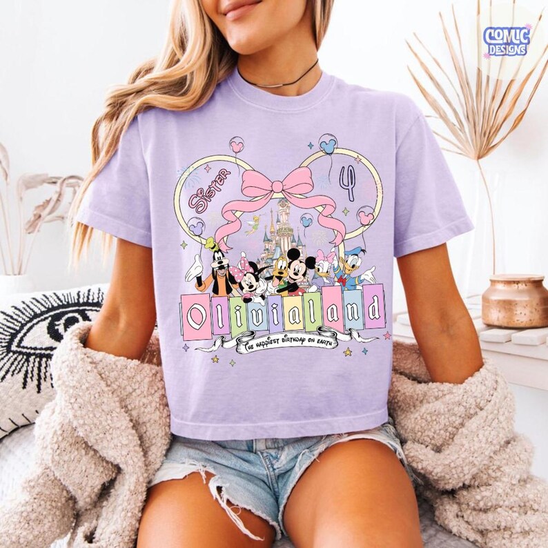 Op de afbeelding: Lichtpaars T-shirt met een Disney-thema. Het ontwerp toont Mickey en Minnie Mouse, Goofy, Donald en Daisy Duck, en de tekst "OliviaLand" en "The happiest birthday on earth."
