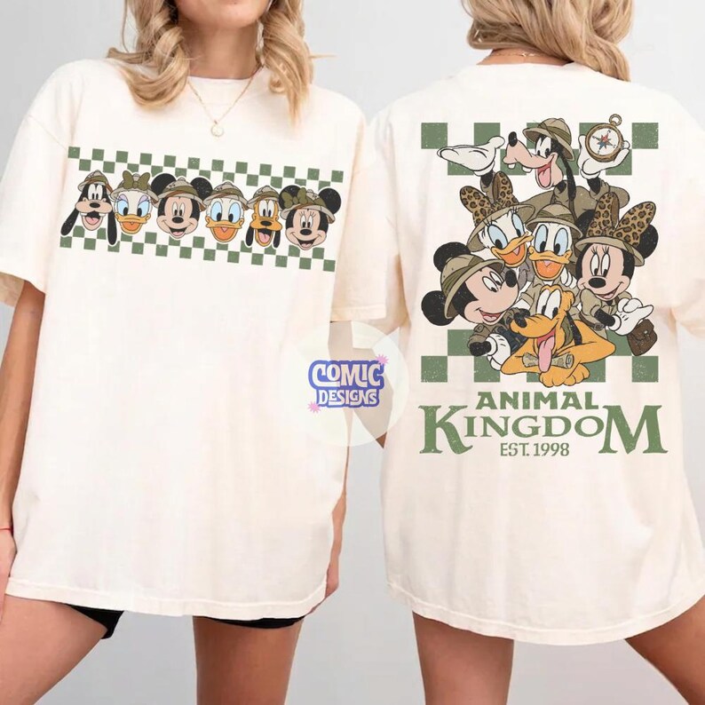Pode incluir: Camiseta creme com personagens da Disney usando chap&eacute;us de safari. A frente exibe uma fileira de rostos de personagens acima de um padr&atilde;o xadrez verde e branco. A parte de tr&aacute;s mostra um grupo de personagens com o texto "ANIMAL KINGDOM EST. 1998".