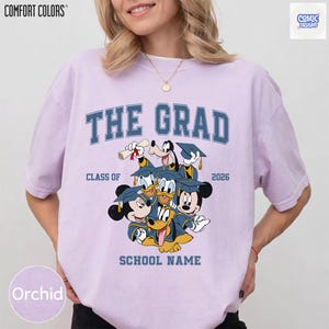 Puede incluir: Camiseta color orqu&iacute;dea con el texto "THE GRAD" en azul, con personajes de Disney con atuendo de graduaci&oacute;n. La camiseta incluye el texto "CLASS OF 2026" y "SCHOOL NAME". La camiseta es de Comfort Colors.