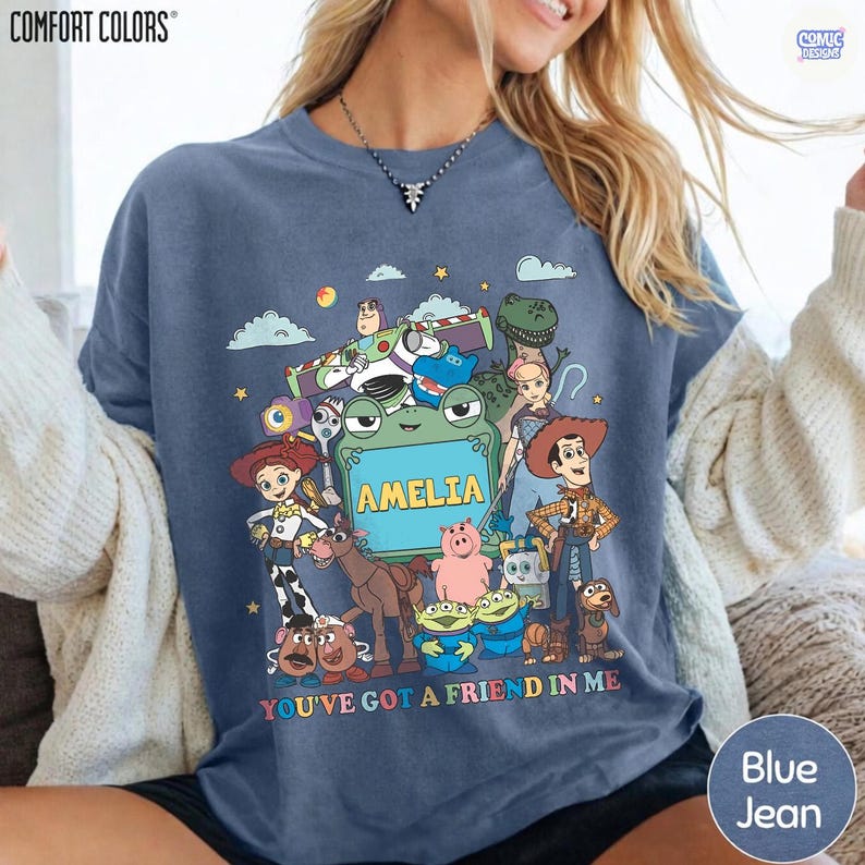 May include: A blue t-shirt featuring a graphic print of various Toy Story characters surrounding a central green frame with the name AMELIA printed in yellow. The text YOU'VE GOT A FRIEND IN ME is printed in colorful letters at the bottom.
