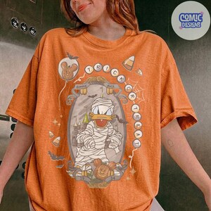 Puede incluir: Una camiseta naranja con un gr&aacute;fico de Donald Duck vestido de momia dentro de un marco ornamentado. El dise&ntilde;o incluye el texto "TRICK OR TREAT" en disposici&oacute;n circular, junto con ilustraciones de murci&eacute;lagos, dulces y un castillo.
