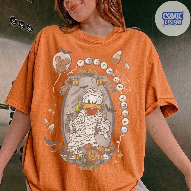 Puede incluir: Una camiseta naranja con un gr&aacute;fico de Donald Duck vestido de momia dentro de un marco ornamentado. El dise&ntilde;o incluye el texto "TRICK OR TREAT" en disposici&oacute;n circular, junto con ilustraciones de murci&eacute;lagos, dulces y un castillo.