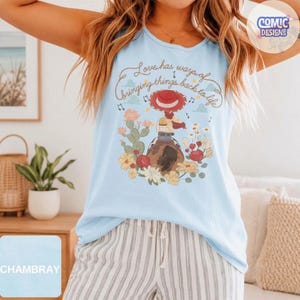 Op de afbeelding: Een lichtblauw mouwloos shirt met een afbeelding van een cowgirl op een paard, omringd door cactussen en bloemen. De tekst luidt, Love has ways of bringing things back to life. Het ontwerp bevat muzieknoten en kleine wolken.