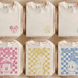 May include: Off-white t-shirts with Disney character designs. The shirts feature Mickey Mouse, Pluto, and Donald Duck, with text such as "Minnie Mouse" and "Pluto" on checkered patterns. They are displayed on woven mats.