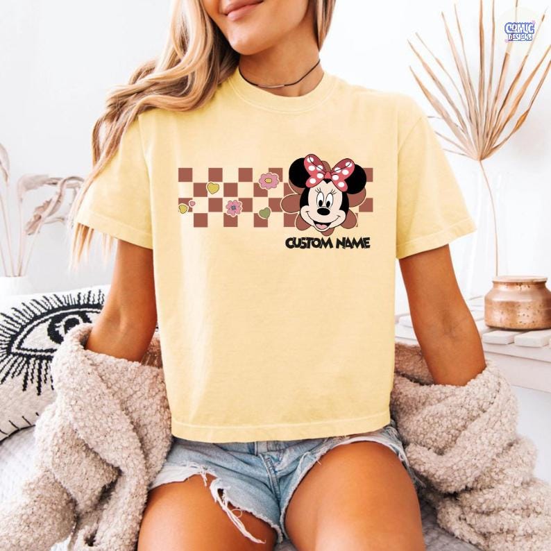 May include: Yellow t-shirt featuring a Minnie Mouse graphic with a red bow and the text "CUSTOM NAME". The design includes a brown and white checkered pattern with floral and heart accents. The shirt has short sleeves.