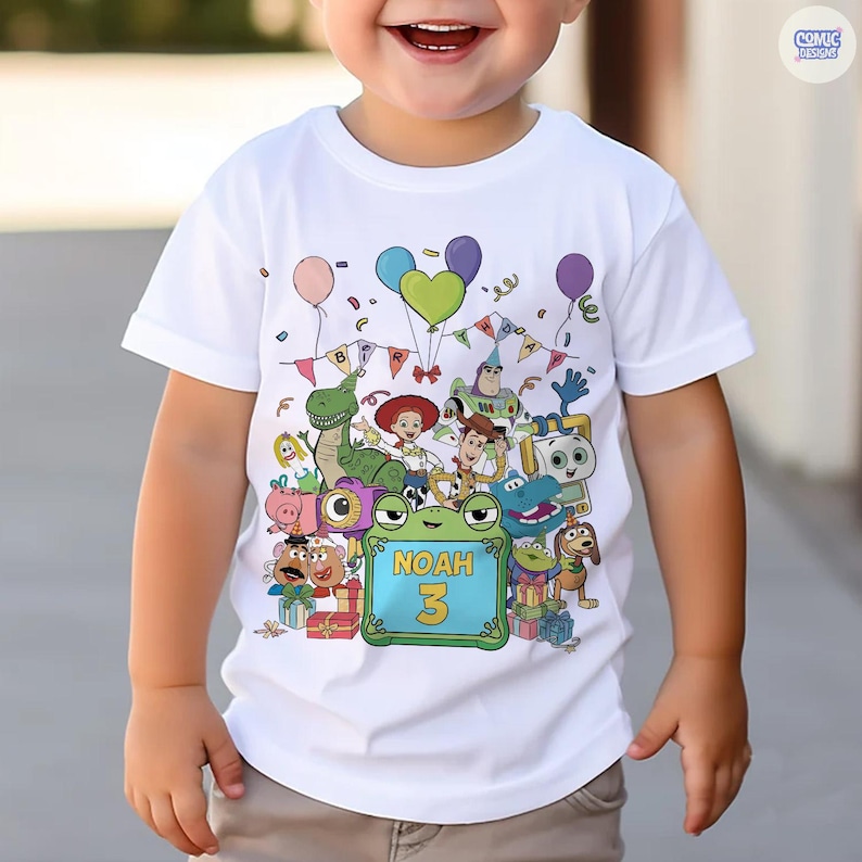 May include: A white short-sleeved t-shirt featuring a colorful graphic of various animated toy characters, balloons, and birthday bunting. A central green square frame contains the text NOAH and the number 3 in bold yellow lettering.