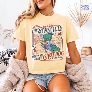 May include: Yellow t-shirt with a graphic of a green dinosaur wearing a hat and suit, holding a hot dog. Text reads "You look like the 4th of July makes me want a hot dog real bad."