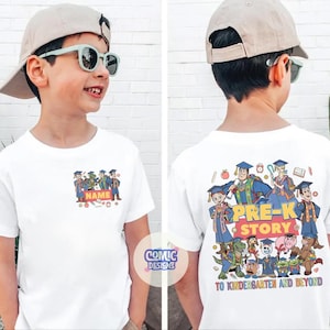 May include: White t-shirt featuring a colorful cartoon design with the text "NAME" on the front and "PRE-K STORY" on the back. The shirt has a crew neck and short sleeves. The design includes graduation caps and characters.