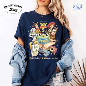 May include: A navy blue t-shirt featuring a Toy Story 5 graphic with characters including Woody, Buzz Lightyear, Jessie, Bullseye, and a green frog in a box labeled Jason. The text reads Toy Story 5 and You've Got A Friend In Me.