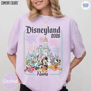Op de afbeelding: Orchidee-kleurig T-shirt met een Disneyland 2026-afbeelding met tekenfilmfiguren en een kasteel. Het ontwerp bevat vuurwerk en het woord "Name" eronder.