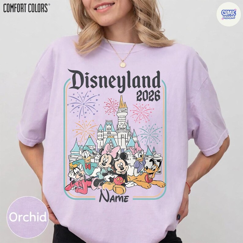 Op de afbeelding: Orchidee-kleurig T-shirt met een Disneyland 2026-afbeelding met tekenfilmfiguren en een kasteel. Het ontwerp bevat vuurwerk en het woord "Name" eronder.