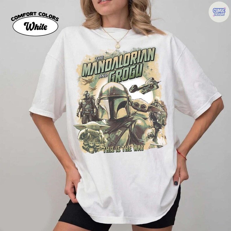 May include: White Comfort Colors t-shirt featuring a graphic of The Mandalorian and Grogu. The design includes the text "The Mandalorian and Grogu" and "This is the Way", along with images of characters and spacecraft.