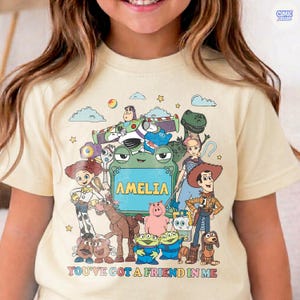 May include: A cream-colored t-shirt featuring a colorful graphic print of various animated characters from the Toy Story franchise. The center of the design includes a green frog-shaped frame with the name AMELIA printed in yellow capital letters.