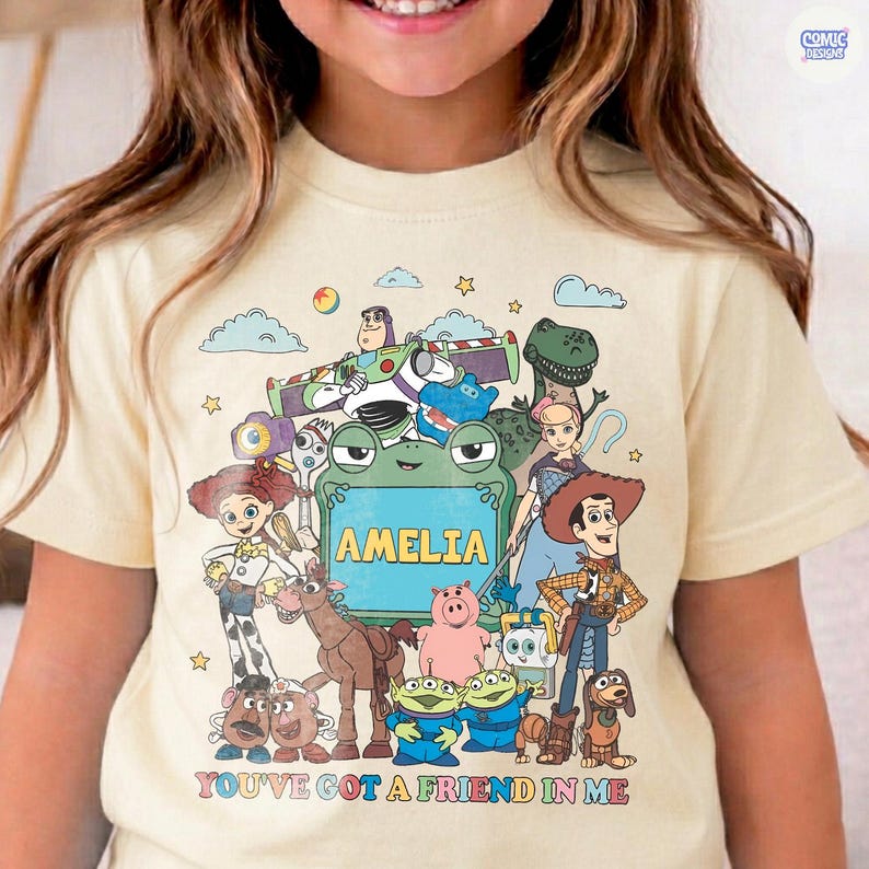 May include: A cream-colored t-shirt featuring a colorful graphic print of various animated characters from the Toy Story franchise. The center of the design includes a green frog-shaped frame with the name AMELIA printed in yellow capital letters.