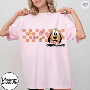May include: A light pink t-shirt featuring a cartoon Pluto graphic with the text "CUSTOM NAME". The design includes a checkered pattern with flowers and hearts. The shirt has short sleeves and a relaxed fit.