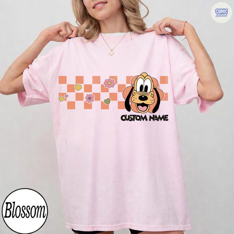 May include: A light pink t-shirt featuring a cartoon Pluto graphic with the text "CUSTOM NAME". The design includes a checkered pattern with flowers and hearts. The shirt has short sleeves and a relaxed fit.