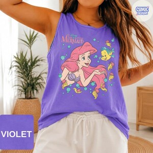 May include: A violet tank top featuring a graphic print of Ariel from The Little Mermaid with long red hair, a purple seashell top, and the text The Little Mermaid. The design includes yellow fish, green bubbles, and small sea creatures on the front.