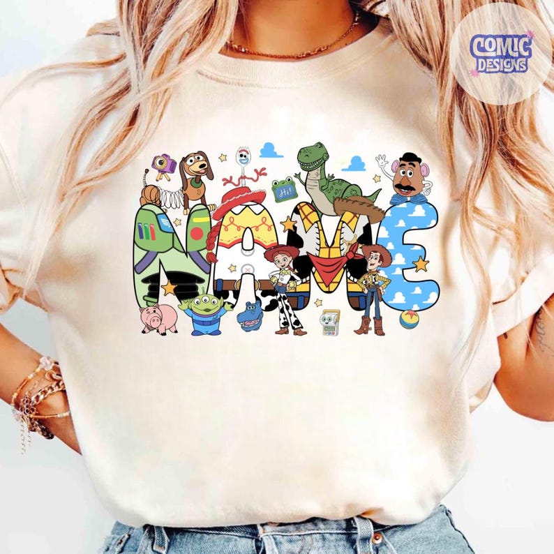 May include: A cream-coloured t-shirt featuring a large graphic print of the word NAME, where each letter is filled with patterns inspired by Toy Story characters. The design includes illustrations of Woody, Buzz Lightyear, Jessie, Rex, and other characters.