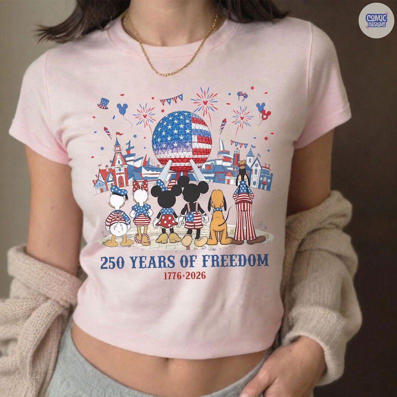 May include: A light pink t-shirt featuring a graphic of cartoon characters standing before a geodesic dome and fireworks. The text reads 250 YEARS OF FREEDOM 1776-2026. The characters are dressed in red, white, and blue patterns with stars and stripes.