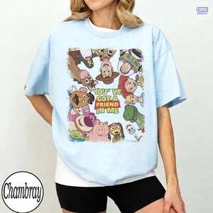 May include: Light blue t-shirt featuring a colorful graphic of characters from the animated film "Toy Story." The graphic includes the text "You've Got a Friend in Me." The shirt is a casual, short-sleeved style.