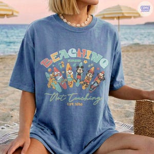 May include: Blue t-shirt with the words "BEACHING" and "Not Teaching" in a colorful design. The shirt features cartoon characters on surfboards. The text "EST. 2026" is also visible.