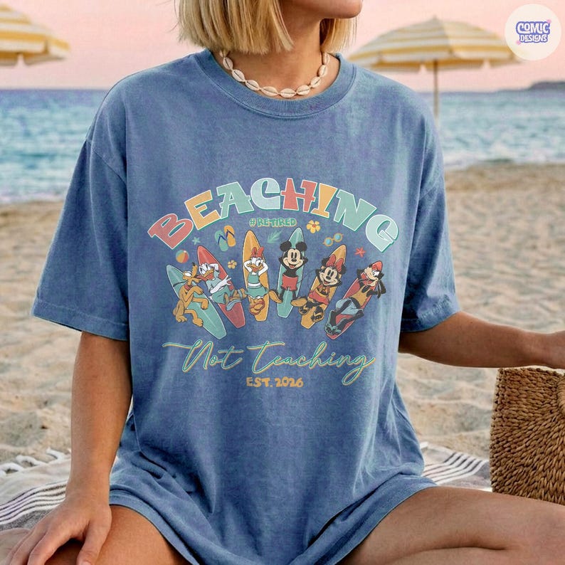 May include: Blue t-shirt with the words "BEACHING" and "Not Teaching" in a colorful design. The shirt features cartoon characters on surfboards. The text "EST. 2026" is also visible.