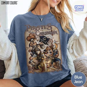 May include: Blue jean-colored sweatshirt featuring a vintage-style graphic of Disney characters dressed as pirates, with the text "Pirates of the Caribbean." The sweatshirt has a crew neck and long sleeves. The brand name "COMFORT COLORS" is visible.