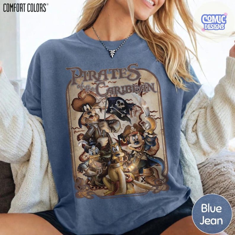 May include: Blue jean-colored sweatshirt featuring a vintage-style graphic of Disney characters dressed as pirates, with the text "Pirates of the Caribbean." The sweatshirt has a crew neck and long sleeves. The brand name "COMFORT COLORS" is visible.