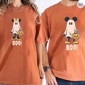 May include: Two orange t-shirts with a Halloween-themed graphic. The graphic features a ghost with Mickey Mouse ears holding a jack-o'-lantern and the word "BOO!". One shirt has a bow on the ghost. The shirts are a relaxed fit.