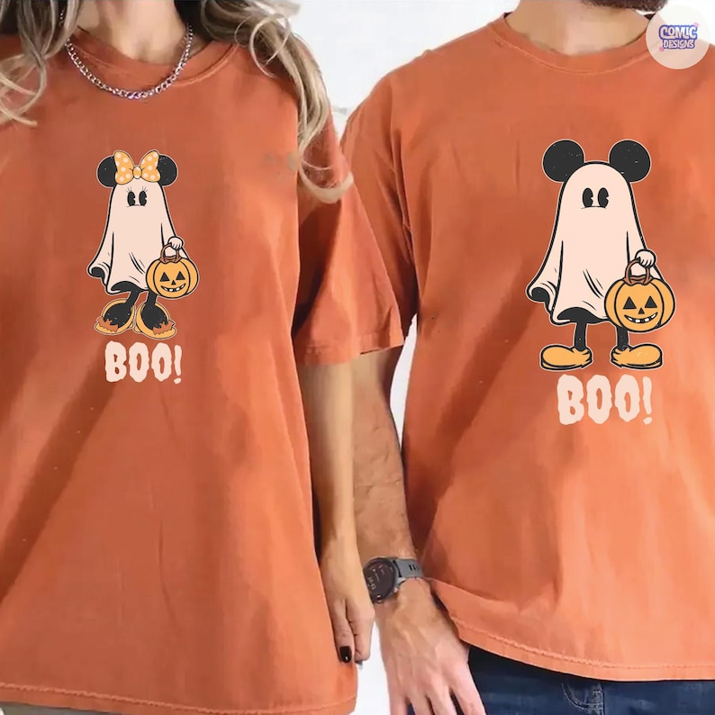 May include: Two orange t-shirts with a Halloween-themed graphic. The graphic features a ghost with Mickey Mouse ears holding a jack-o'-lantern and the word "BOO!". One shirt has a bow on the ghost. The shirts are a relaxed fit.