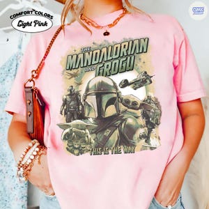 May include: Light pink t-shirt featuring a graphic of The Mandalorian and Grogu, with the text "THIS IS THE WAY". The shirt has the "Comfort Colors" and "Comic Designs" logos.