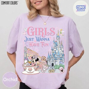 May include: Orchid-colored t-shirt with the text "GIRLS JUST WANNA HAVE FUN" and a graphic featuring Disney characters, a castle, and other theme park elements. The shirt is from Comfort Colors and has a Comic Designs logo.