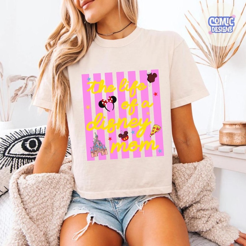 Puede incluir: Camiseta color crema con el texto "The life of a Disney mom" en amarillo, sobre un fondo a rayas rosas y blancas. El dise&ntilde;o incluye iconos de Disney como orejas de Mickey y un castillo.