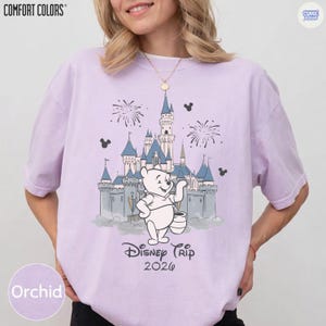 May include: Orchid-colored t-shirt with a graphic of a castle, fireworks, and Winnie the Pooh. The text "Disney Trip 2026" is below the graphic. The shirt has the "Comfort Colors" logo in the upper left corner.