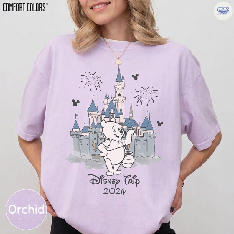 May include: Orchid-colored t-shirt with a graphic of a castle, fireworks, and Winnie the Pooh. The text "Disney Trip 2026" is below the graphic. The shirt has the "Comfort Colors" logo in the upper left corner.