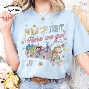 May include: Light blue Comfort Colors t-shirt with the text "HOLD ON TIGHT, Here we go!" and a colorful roller coaster graphic featuring cartoon characters. The shirt has a crew neck and short sleeves.