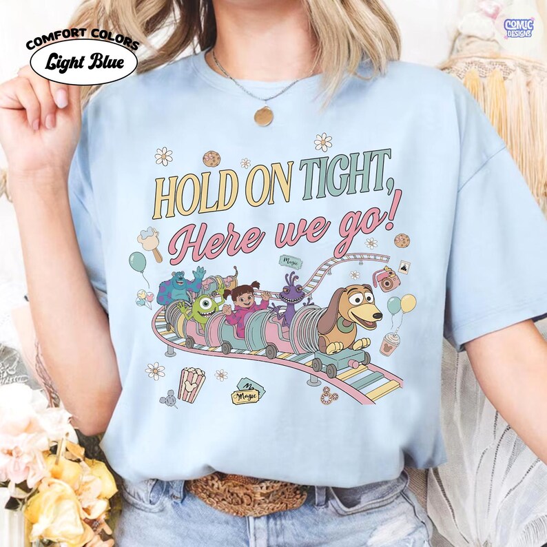 May include: Light blue Comfort Colors t-shirt with the text "HOLD ON TIGHT, Here we go!" and a colorful roller coaster graphic featuring cartoon characters. The shirt has a crew neck and short sleeves.