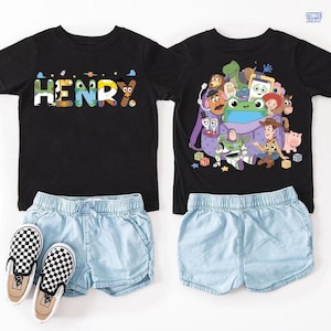 May include: Two black t-shirts and two pairs of light blue shorts. One shirt features the name HENRY in colorful patterned letters. The other shirt displays a graphic of various animated toy characters emerging from a purple backpack. A pair of black and white checkered slip-on shoes is included.