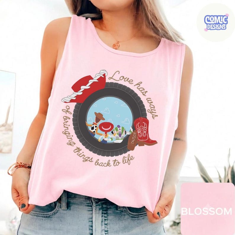 May include: A light pink sleeveless tank top featuring a graphic print of a black tire framing a blue sky background with cartoon characters, a red cowboy hat, and a red cowboy boot. The text reads, Love has ways of bringing things back to life.