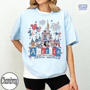 May include: Light blue t-shirt featuring a patriotic design with Disney characters, fireworks, and a castle. The word "America" is prominently displayed, along with the years "1776-2026". The shirt has a crew neck and short sleeves.