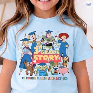 May include: Light blue t-shirt featuring a colorful graphic of animated characters with the text "3rd Grade Story" and "To Fourth Grade and Beyond". The design includes various cartoon figures in graduation attire.