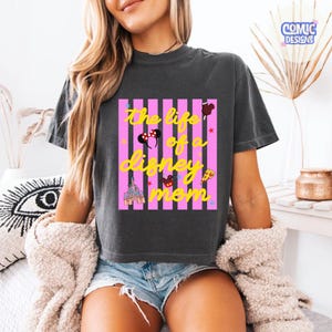 Puede incluir: Camiseta gris oscuro con el texto amarillo "The life of a Disney mom" sobre rayas verticales rosas. El dise&ntilde;o incluye iconos de Disney como orejas de Mickey Mouse, un castillo y pizza.