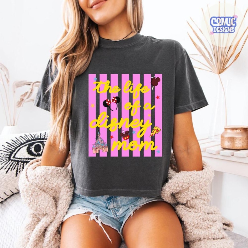 Puede incluir: Camiseta gris oscuro con el texto amarillo "The life of a Disney mom" sobre rayas verticales rosas. El dise&ntilde;o incluye iconos de Disney como orejas de Mickey Mouse, un castillo y pizza.