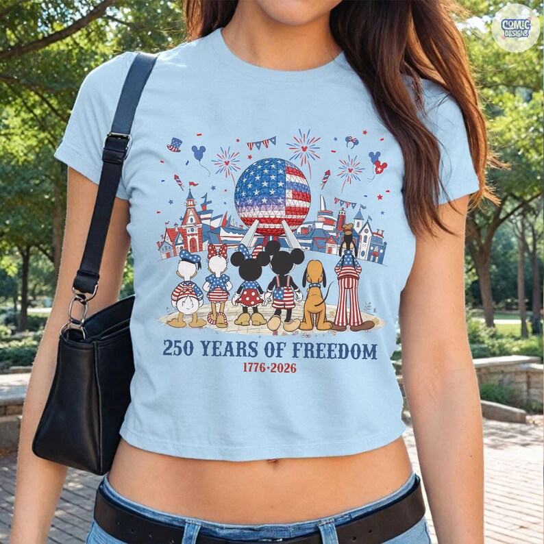 May include: A light blue t-shirt featuring a graphic of cartoon characters standing before a geodesic dome and castle. The design includes fireworks, stars, and stripes, with the text 250 YEARS OF FREEDOM 1776-2026 printed in blue and red.