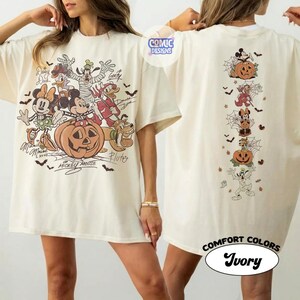 Op de afbeelding: Een ivoorkleurig t-shirt met een grote Halloween-afbeelding op de voorkant met Mickey Mouse, Minnie Mouse, Goofy, Donald Duck en Pluto rond een pompoen. De achterkant toont een verticale kolom van dezelfde personages met spinnenwebben.