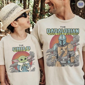 Puede incluir: Camisetas beige con gr&aacute;ficos a juego. Una camiseta presenta "THE CHILD" con un dibujo animado de Grogu, la otra dice "THE DADALORIAN" con una ilustraci&oacute;n de Mandaloriano. Ambos dise&ntilde;os incluyen un sol rojo y humo.