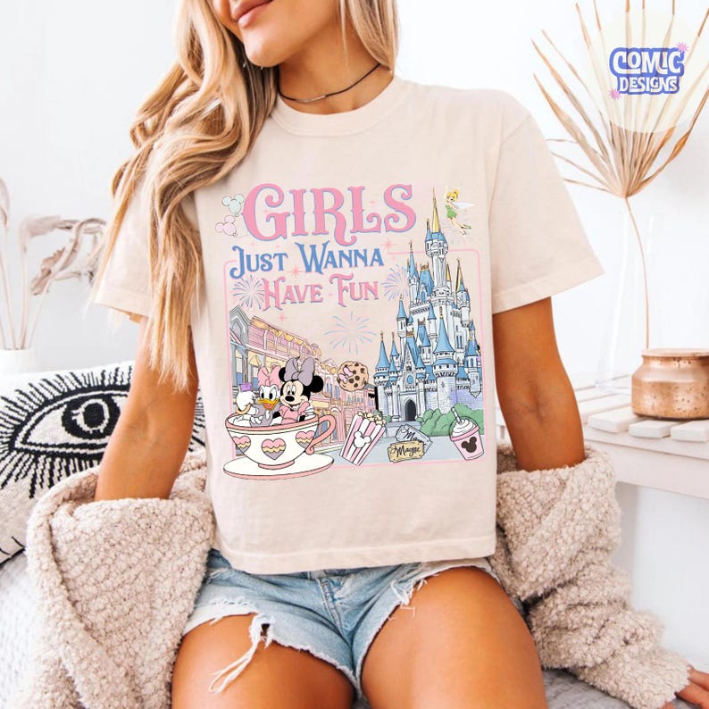 May include: Cream-colored t-shirt with a colorful graphic featuring the text "GIRLS JUST WANNA HAVE FUN". The graphic includes Disney characters, a castle, and other theme park elements. The shirt has short sleeves.