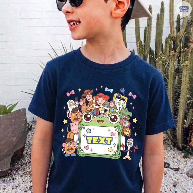 May include: A navy blue t-shirt featuring a colorful graphic print of various Toy Story characters surrounding a central light green frog-shaped frame with the word TEXT printed in bold blue letters, accented by small stars and bows.